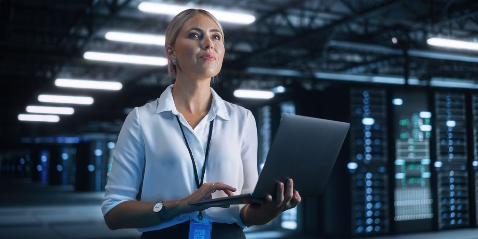 female data center IT technician standing at the server rack corridor with laptop computer